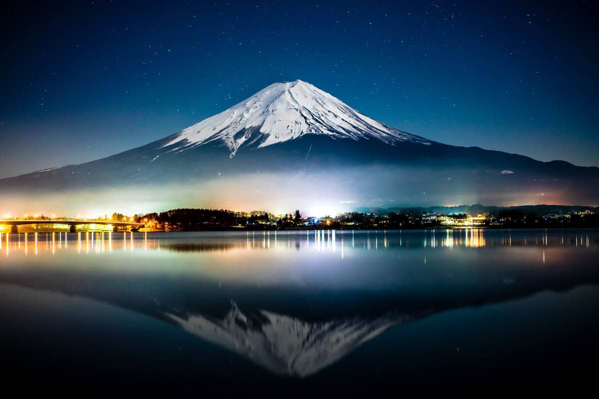 Snowcapped Mount Fuji reflects across dark water beside lakeside lights in Japan.