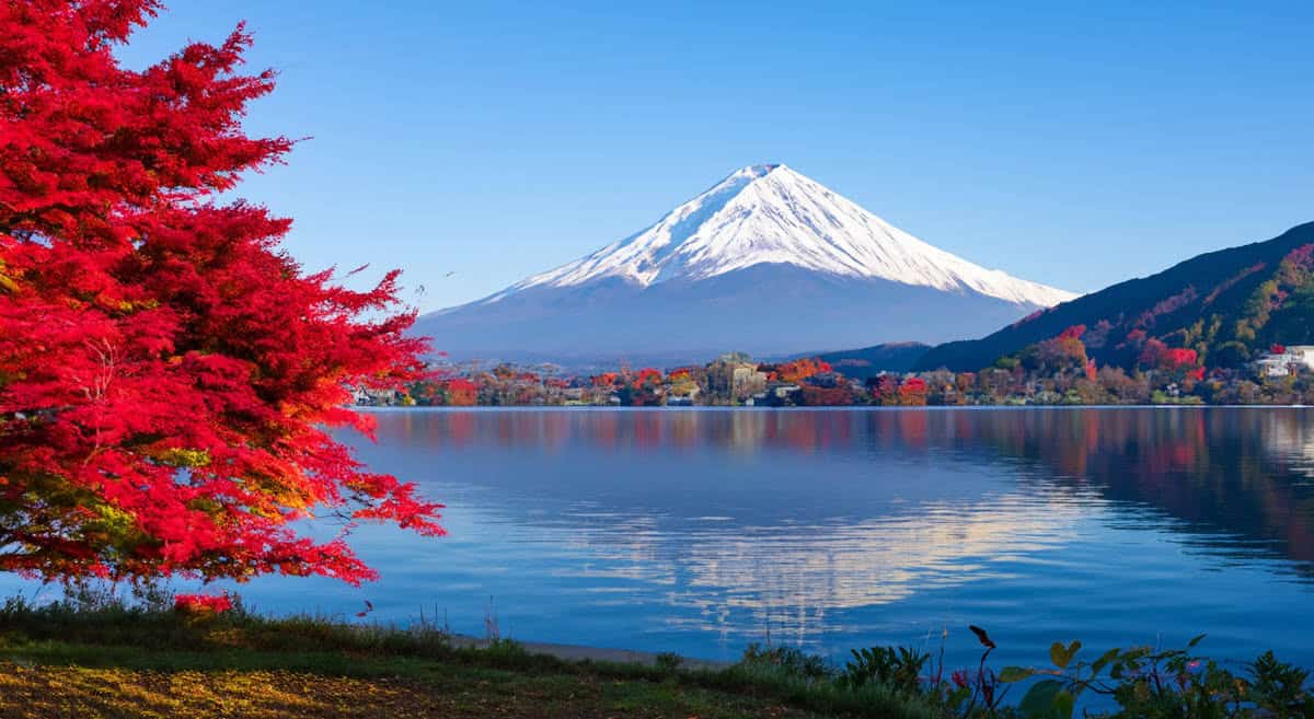 Crimson trees border a blue lake below snowcapped Mount Fuji in Japan.