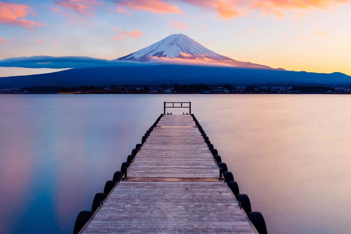 A long wooden pier leads over still water toward Mount Fuji at dusk in Japan.
