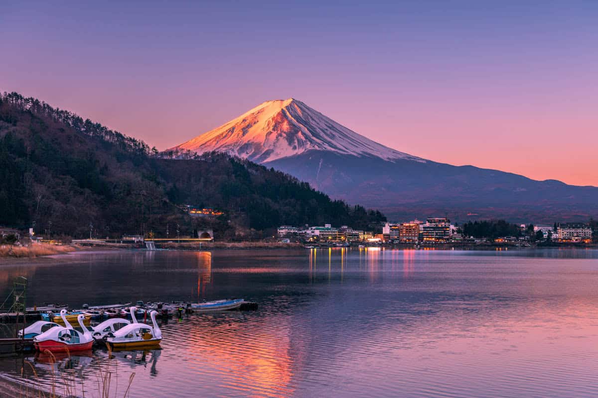 Moored boats line a quiet lakeshore beneath sunset-tinted Mount Fuji in Japan.