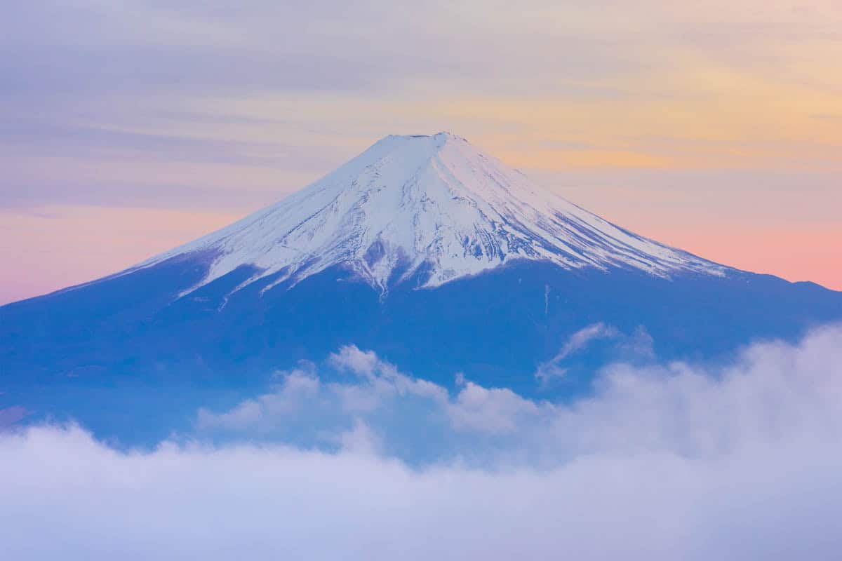 Snow-covered Mount Fuji rises above layered clouds beneath a pastel dawn sky in Japan.